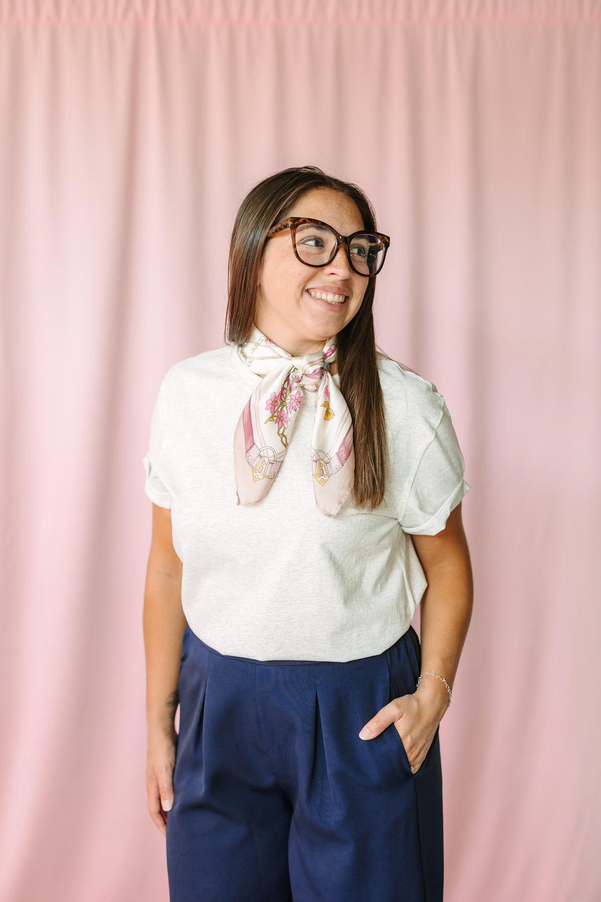 Woman wearing heather gray short-sleeve tee with cream printed neck scarf and navy wide-leg trousers, smiling against pink curtain backdrop