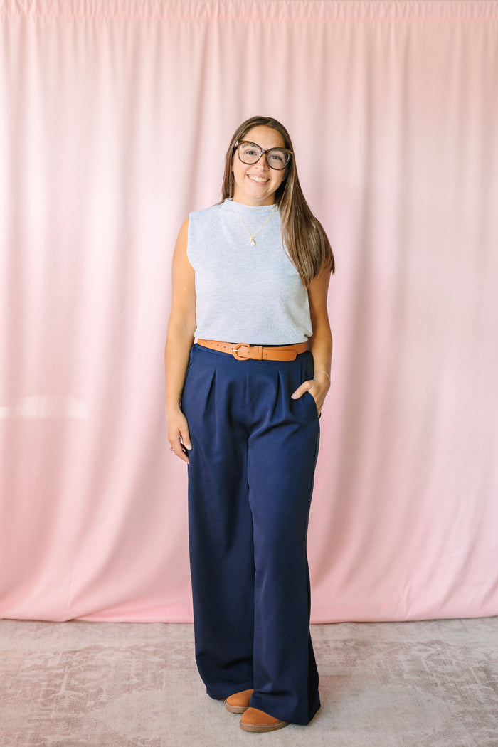 Navy wide leg dress pants styled with a light gray sleeveless top and tan belt on a woman with glasses, pink backdrop