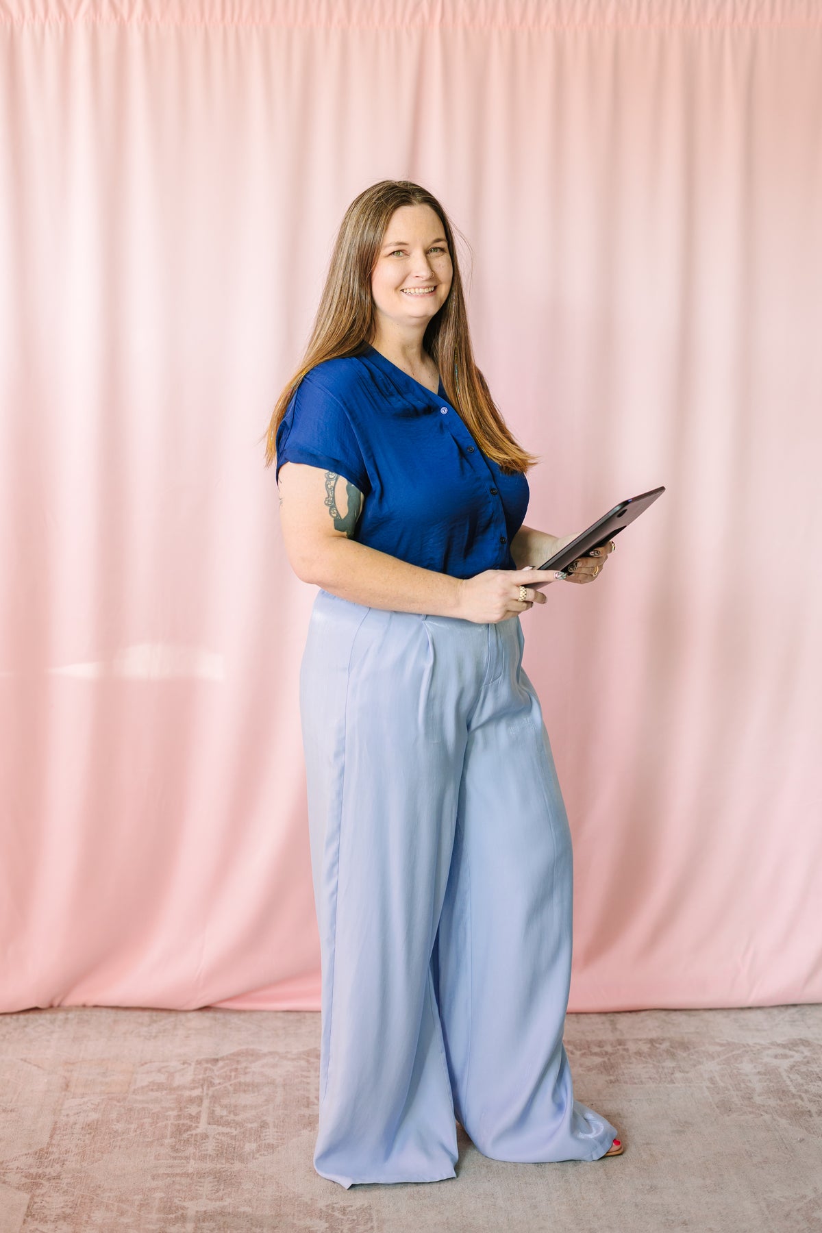Woman wearing a navy blue short-sleeve button-front blouse tucked into light blue wide-leg trousers, holding a tablet, smiling in a side profile pose against a pink draped backdrop