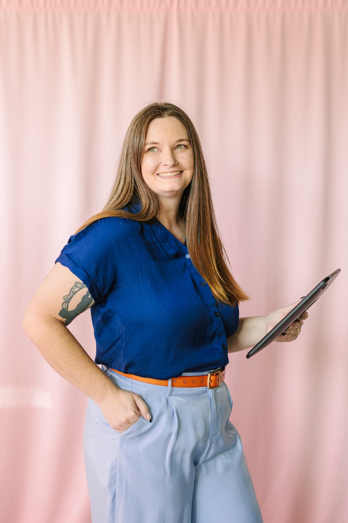 Woman wearing a navy short-sleeve button-down blouse with light blue wide-leg trousers and brown leather belt, holding a tablet, smiling against a pink backdrop
