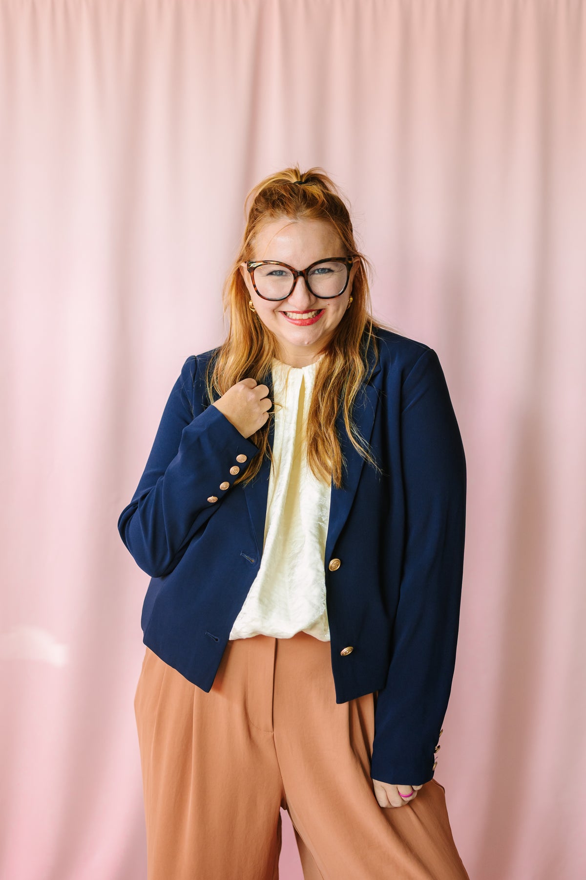 Woman smiling in navy blazer with gold buttons over cream blouse and camel wide-leg trousers, pink backdrop