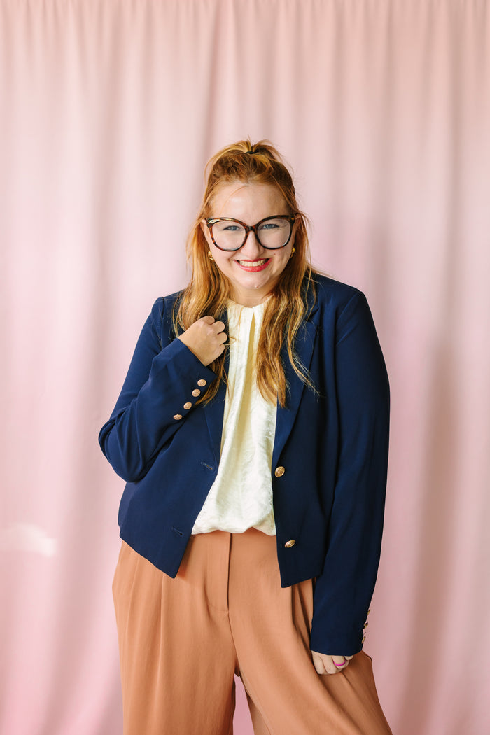 Woman smiling in navy blazer with gold buttons over cream blouse and camel wide-leg trousers, pink backdrop