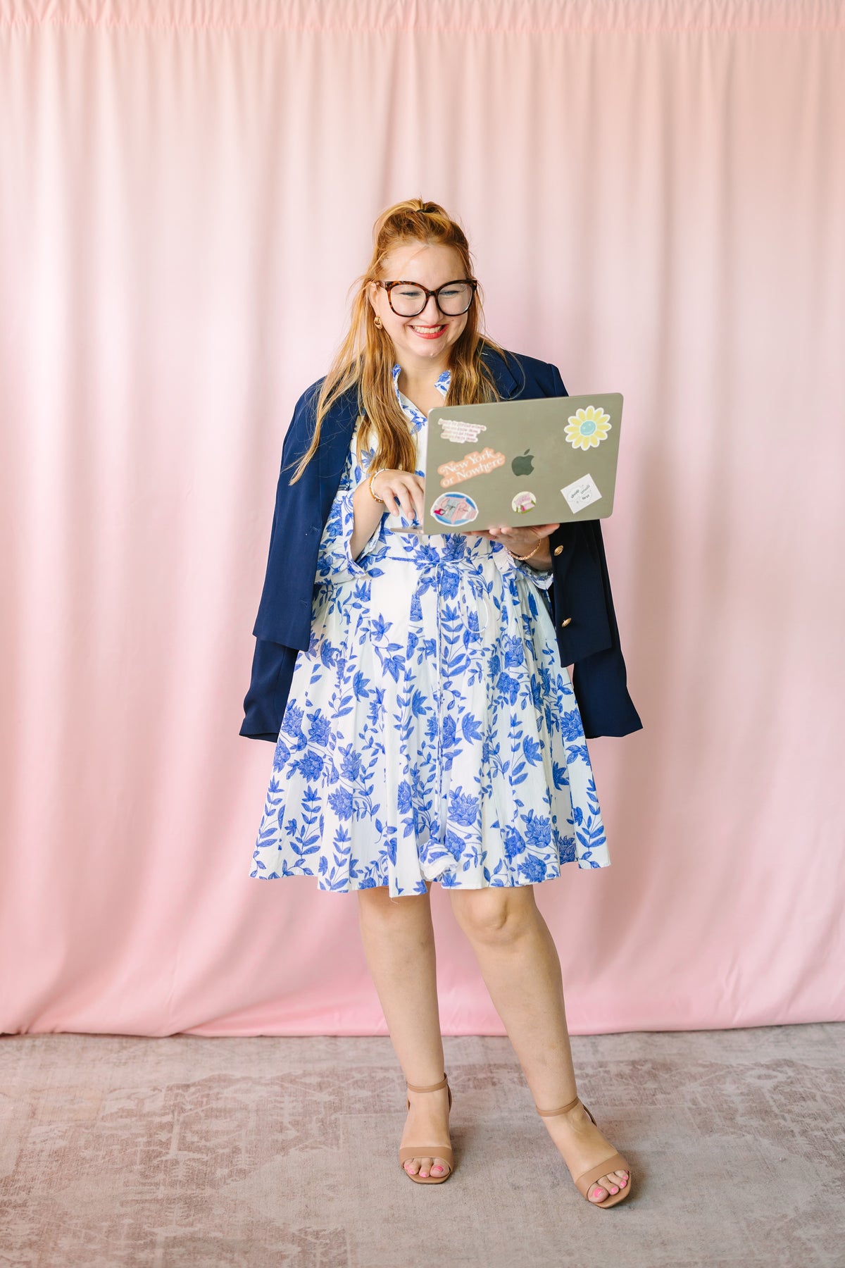 Woman in navy blazer over blue and white floral dress holding a laptop, full-length shot on pink backdrop