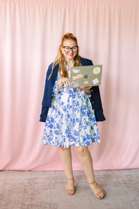 Woman in navy blazer over blue and white floral dress holding a laptop, full-length shot on pink backdrop