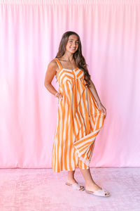 Model wearing an orange and cream striped midi dress with scoop neckline, wide straps, empire waist, and side pockets, styled with white slide sandals against a pink backdrop.

