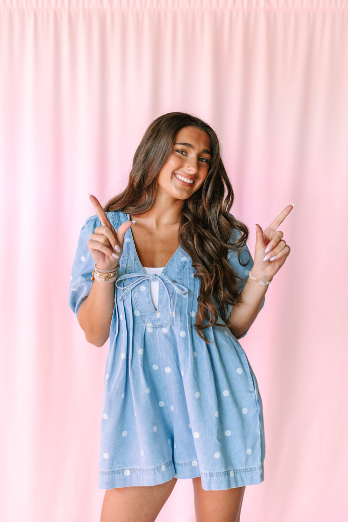 A woman posing in a light blue denim romper with white polka dots against a pink curtain backdrop.