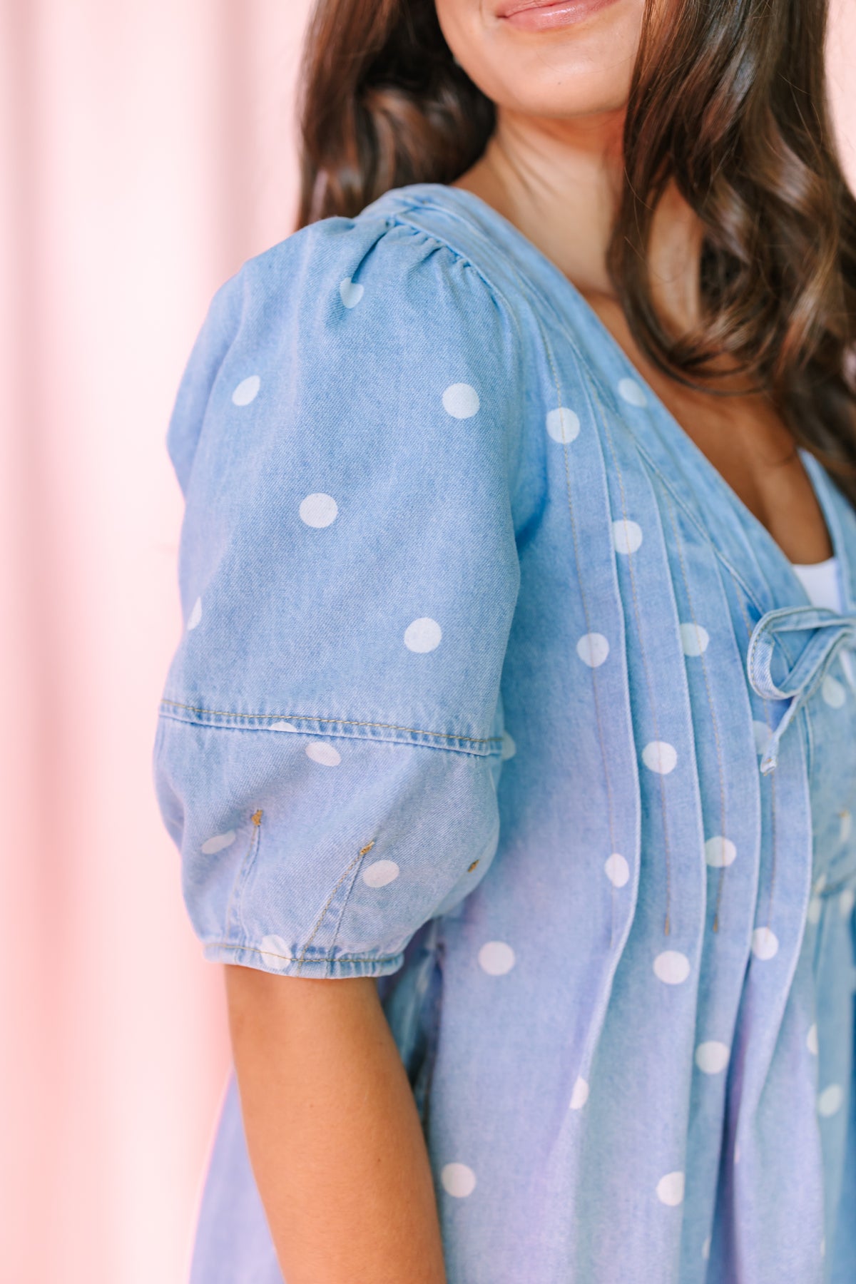 A close-up of a woman wearing a light blue denim romper with white polka dots, featuring puff sleeves and a V-neckline, set against a soft pink background.