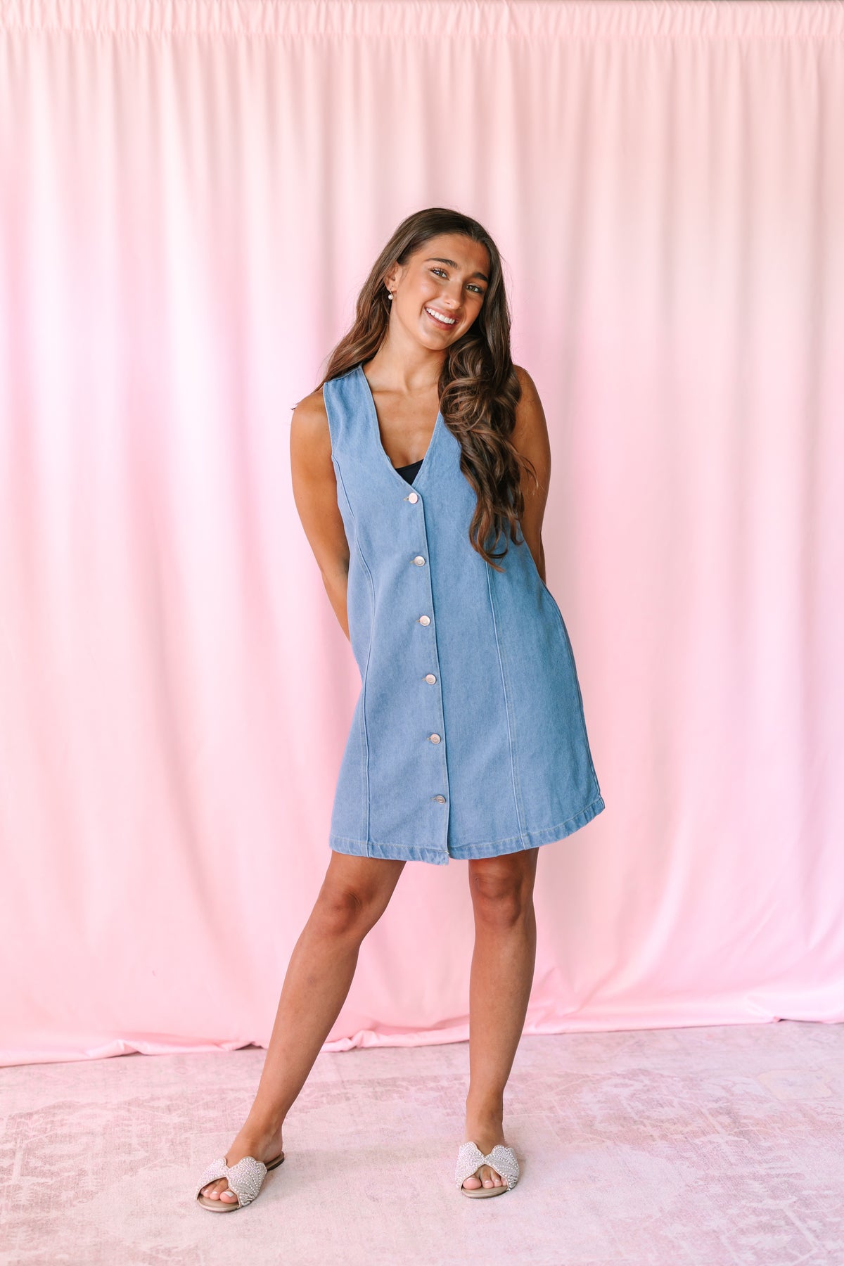 A woman modeling a sleeveless light blue denim dress with a button-down front, standing against a pink fabric backdrop.