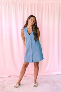 A woman modeling a sleeveless light blue denim dress with a button-down front, standing against a pink fabric backdrop.
