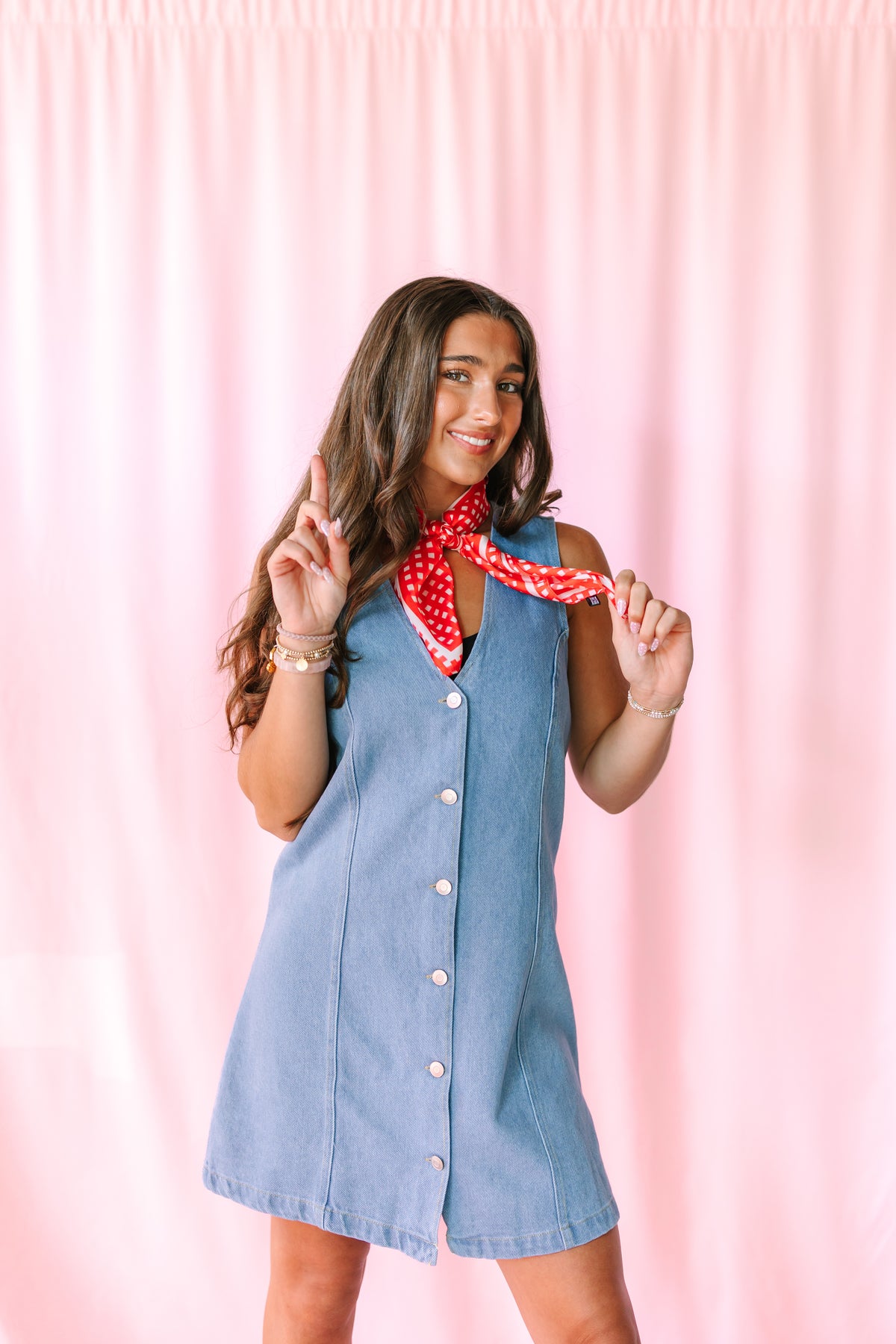 A woman modeling a sleeveless light blue denim dress with a red and white polka dot neck scarf, posing in front of a pink curtain backdrop.