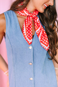 A close-up lifestyle shot of a woman wearing a sleeveless light blue denim button down dress with a red and white patterned neck scarf, set against a solid pink background.