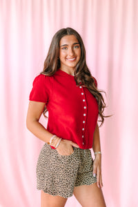 A woman posing in a vibrant red short-sleeve button-up shirt and leopard-print shorts against a pink draped backdrop.