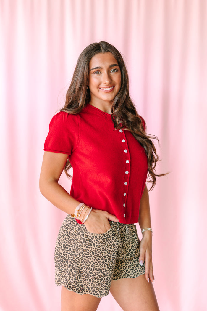 A woman posing in a vibrant red short-sleeve button-up shirt and leopard-print shorts against a pink draped backdrop.