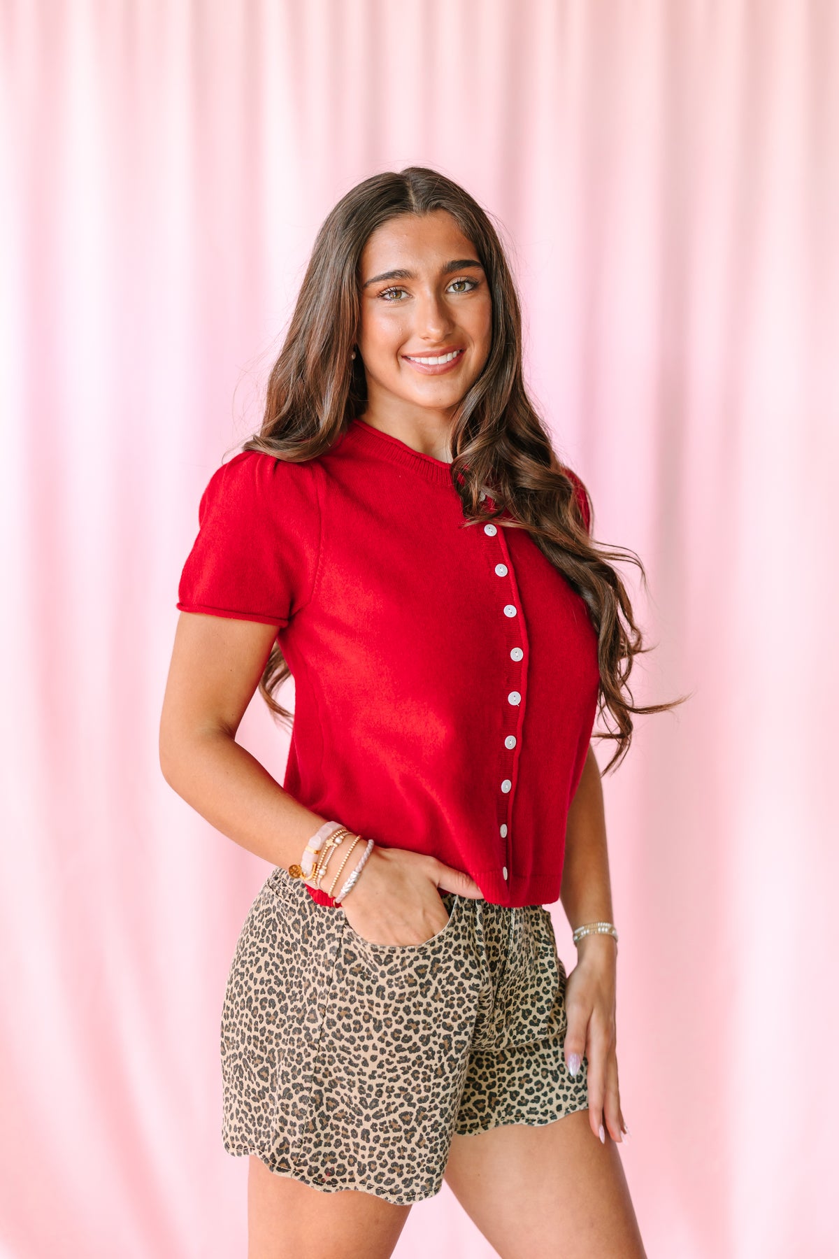 A woman posing in a vibrant red short-sleeve button-up shirt and leopard-print shorts against a pink draped backdrop.