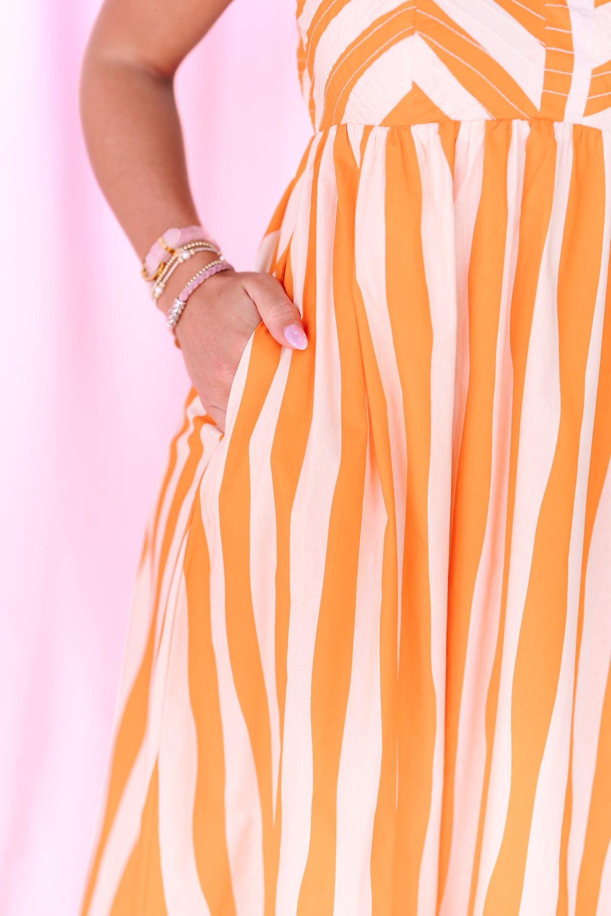 Close-up of a person wearing a sleeveless dress with vertical orange and cream stripes, with one hand in a pocket against a soft pink background.