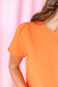 Close-up of a person wearing a bright orange short-sleeve knit top against a soft pink background.