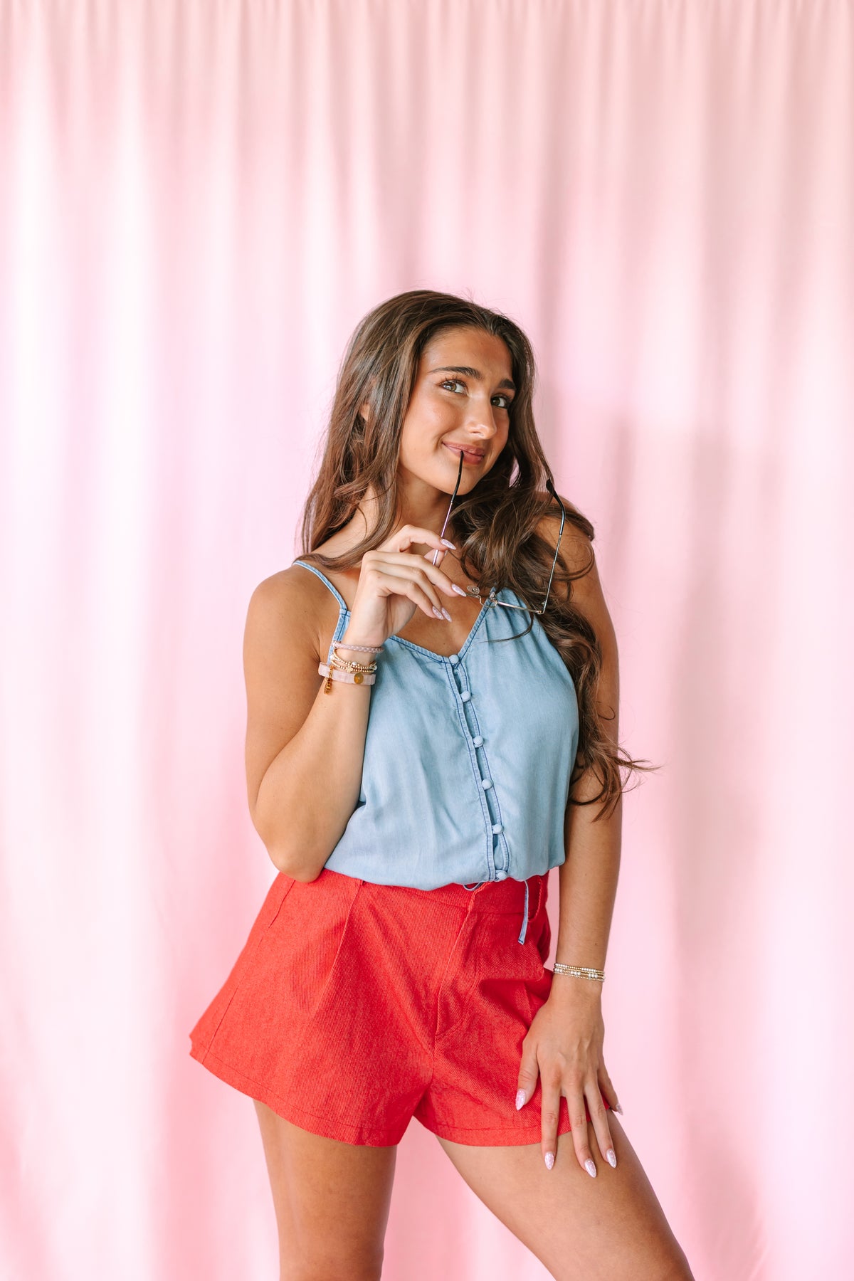 A woman posing in a light blue denim  spaghetti strap top and red shorts against a pink curtain backdrop.