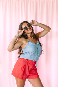 A woman posing in a light blue denim camisole and red shorts against a pink curtain backdrop.