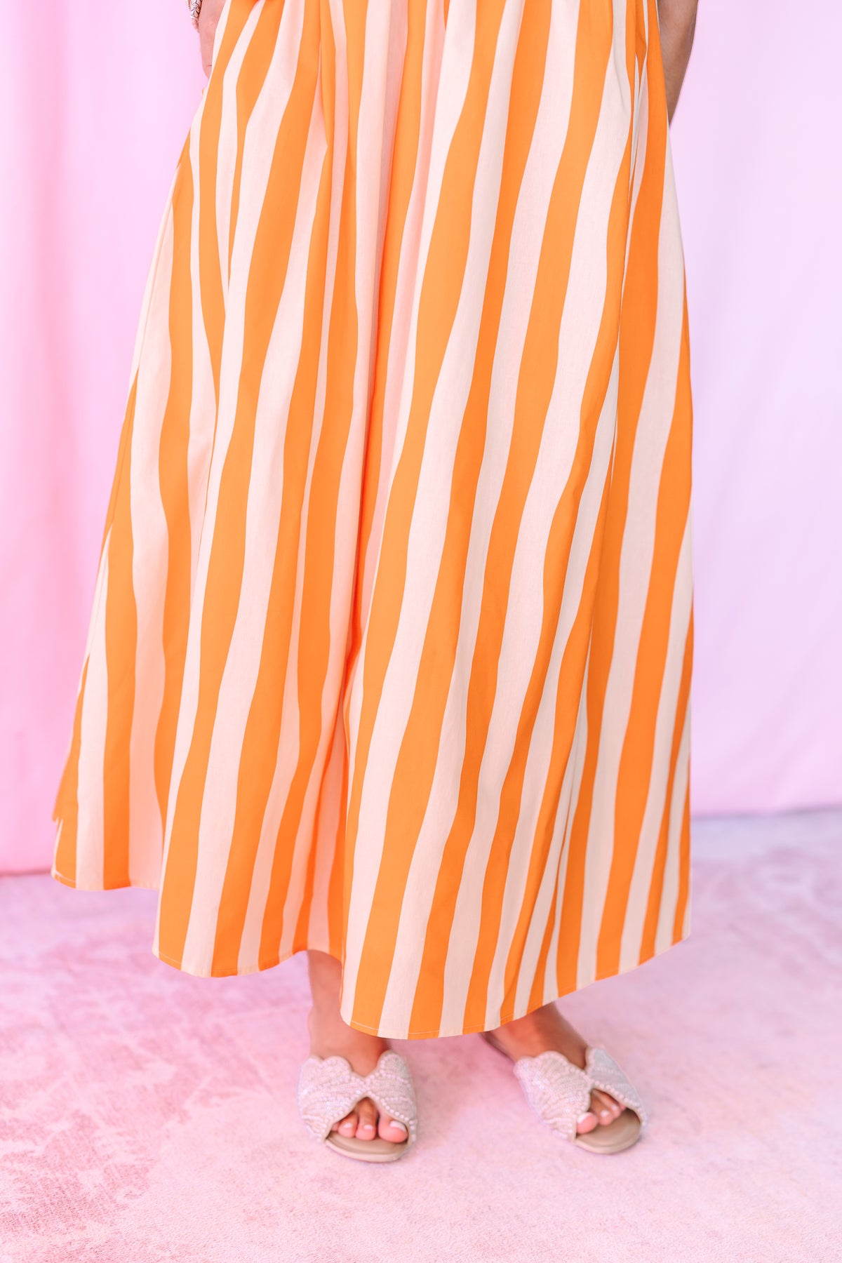 A person wearing a flowing skirt with vertical orange and cream stripes, paired with slide sandals, standing against a soft pink backdrop.