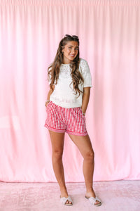A young woman stands against a pink curtain backdrop wearing a white short-sleeve knit top and red-and-white gingham shorts, with white slide sandals.