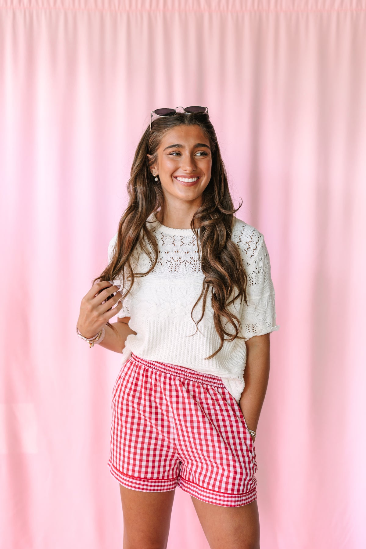 A young woman smiling in front of a pink curtain backdrop, wearing a white short-sleeve knit top with lace detailing and red-and-white gingham shorts.