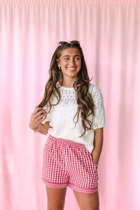 A young woman smiling in front of a pink curtain backdrop, wearing a white short-sleeve knit top with lace detailing and red-and-white gingham shorts.