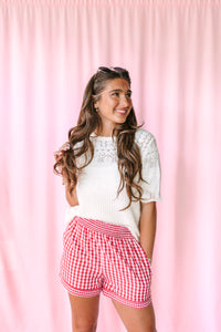 A woman posing in front of a pink curtain backdrop, wearing a white knit top and red and white gingham check shorts.