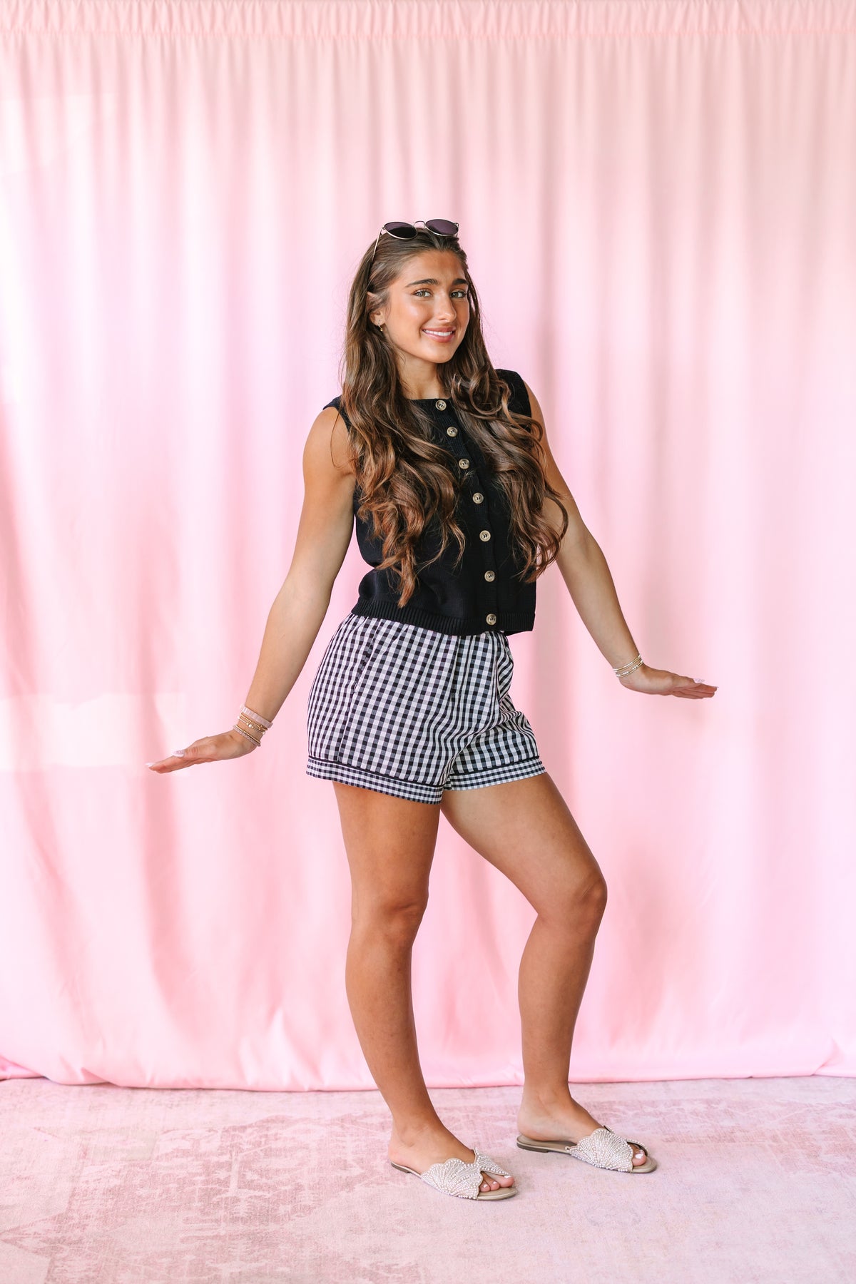 A woman posing in a black sleeveless top with buttons and black-and-white gingham shorts, standing against a pink curtain backdrop.