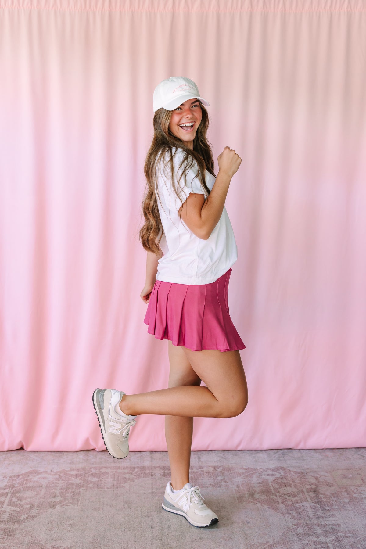 Woman posing playfully in a mauve pleated mini skirt, white t-shirt, white baseball cap, and white sneakers against a pink backdrop
