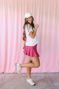 Woman posing playfully in a mauve pleated mini skirt, white t-shirt, white baseball cap, and white sneakers against a pink backdrop