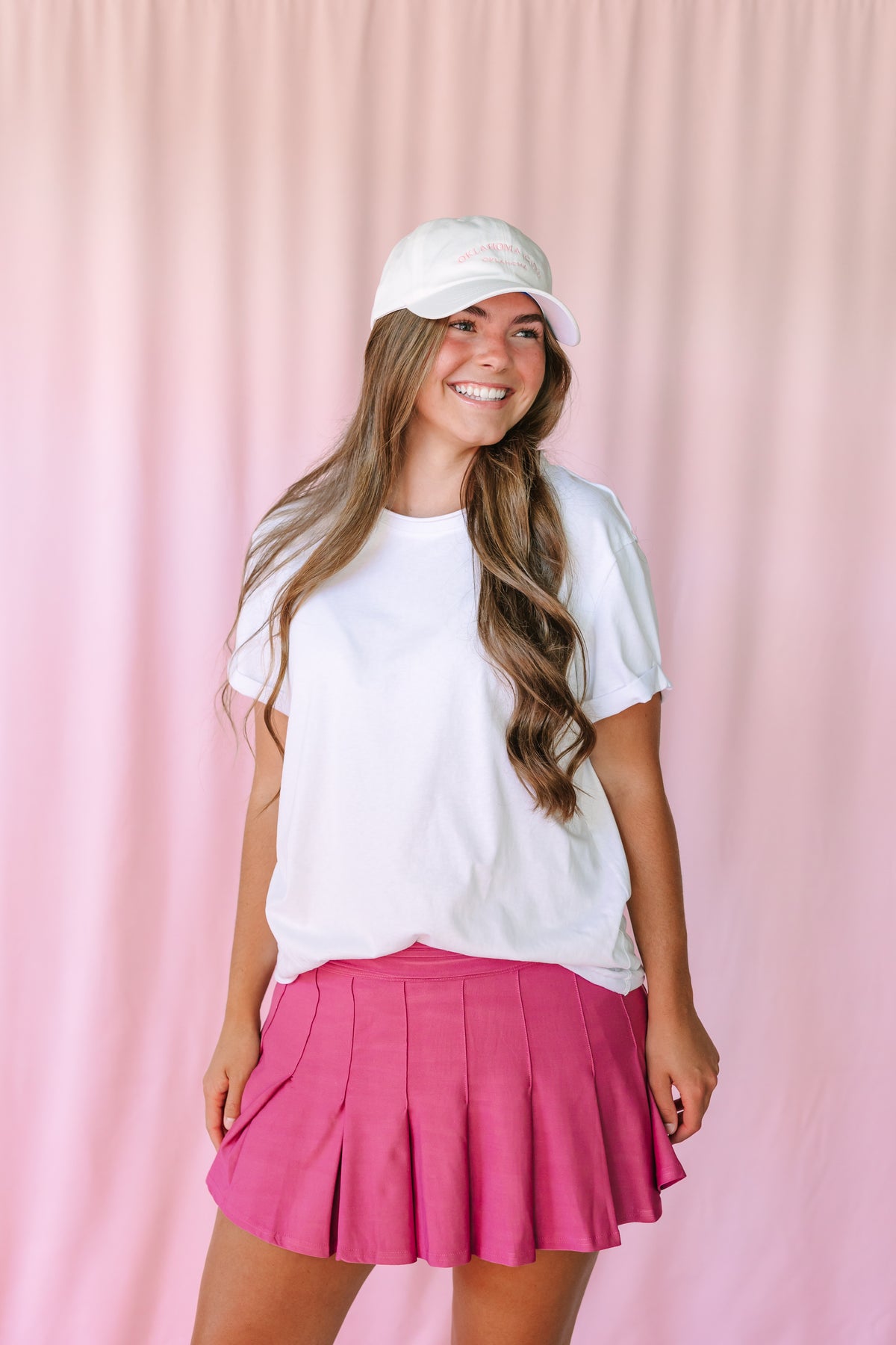 Woman smiling in a mauve pleated mini skirt and white oversized t-shirt with a white baseball cap, pink curtain background