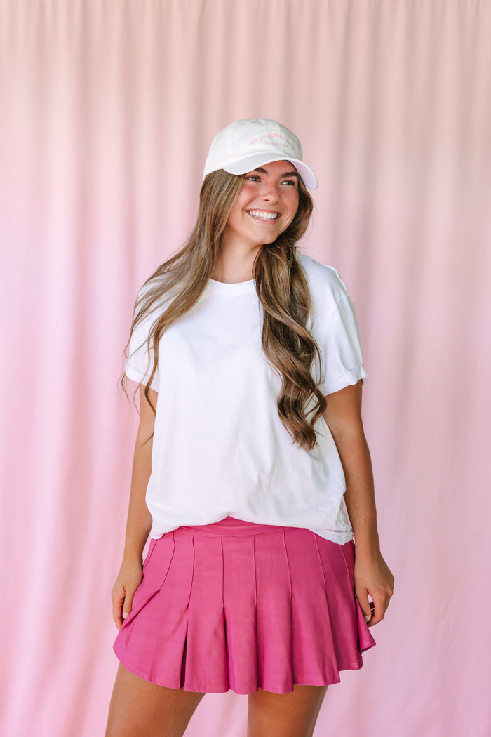 Woman smiling in a mauve pleated mini skirt and white oversized t-shirt with a white baseball cap, pink curtain background