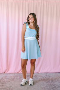 Woman smiling in a light blue sleeveless fit-and-flare mini dress with ruffle shoulder straps, white and green stripe waistband, and white sneakers against a pink backdrop