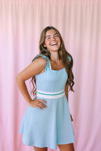 Woman laughing with hand on hip in a light blue sleeveless mini dress with ruffle straps and white and green stripe waistband, pink background