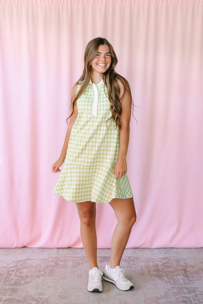 Woman wearing a sage green gingham sleeveless mini dress with white collar, tiered ruffle hem, and white sneakers against a pink background
