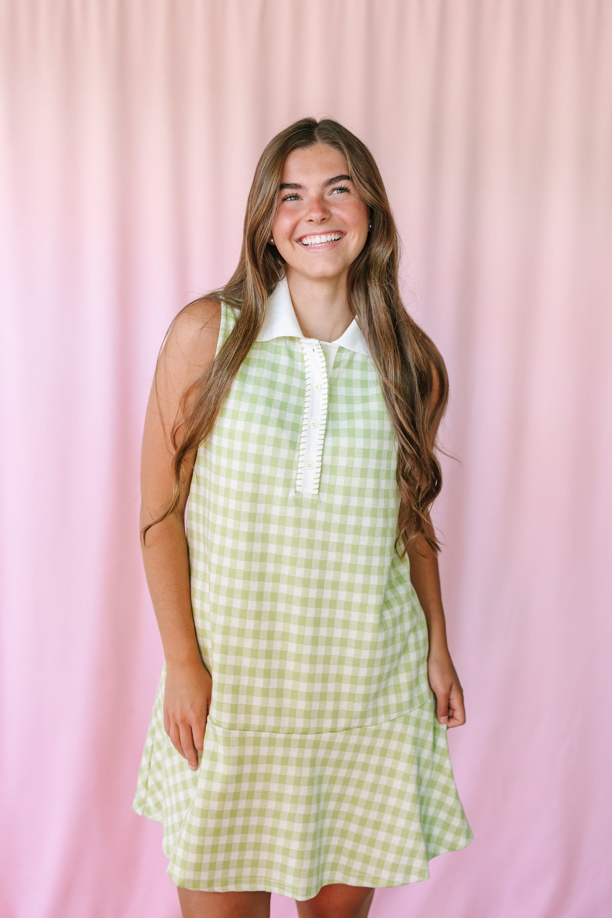 Woman smiling in a sage green gingham sleeveless polo dress with white collar and button front, pink backdrop