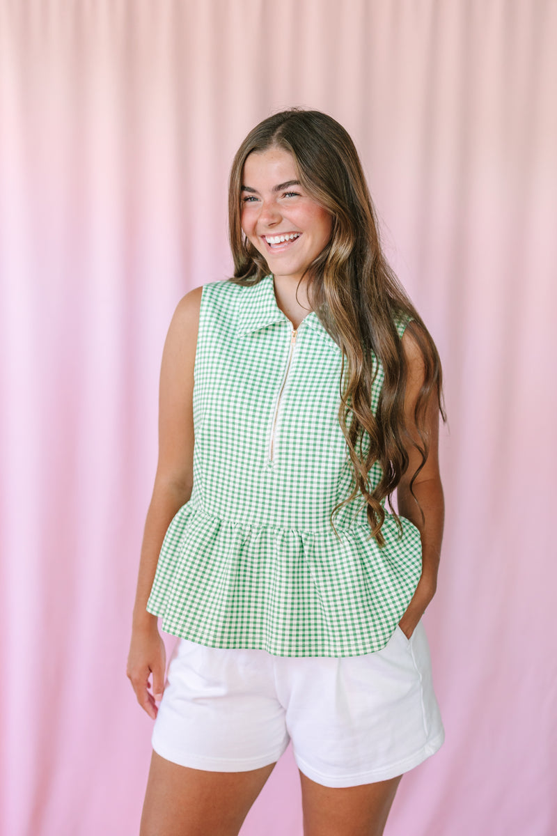 Woman smiling in a green gingham sleeveless peplum top with gold half-zip and collared neckline, styled with white shorts against a pink backdrop