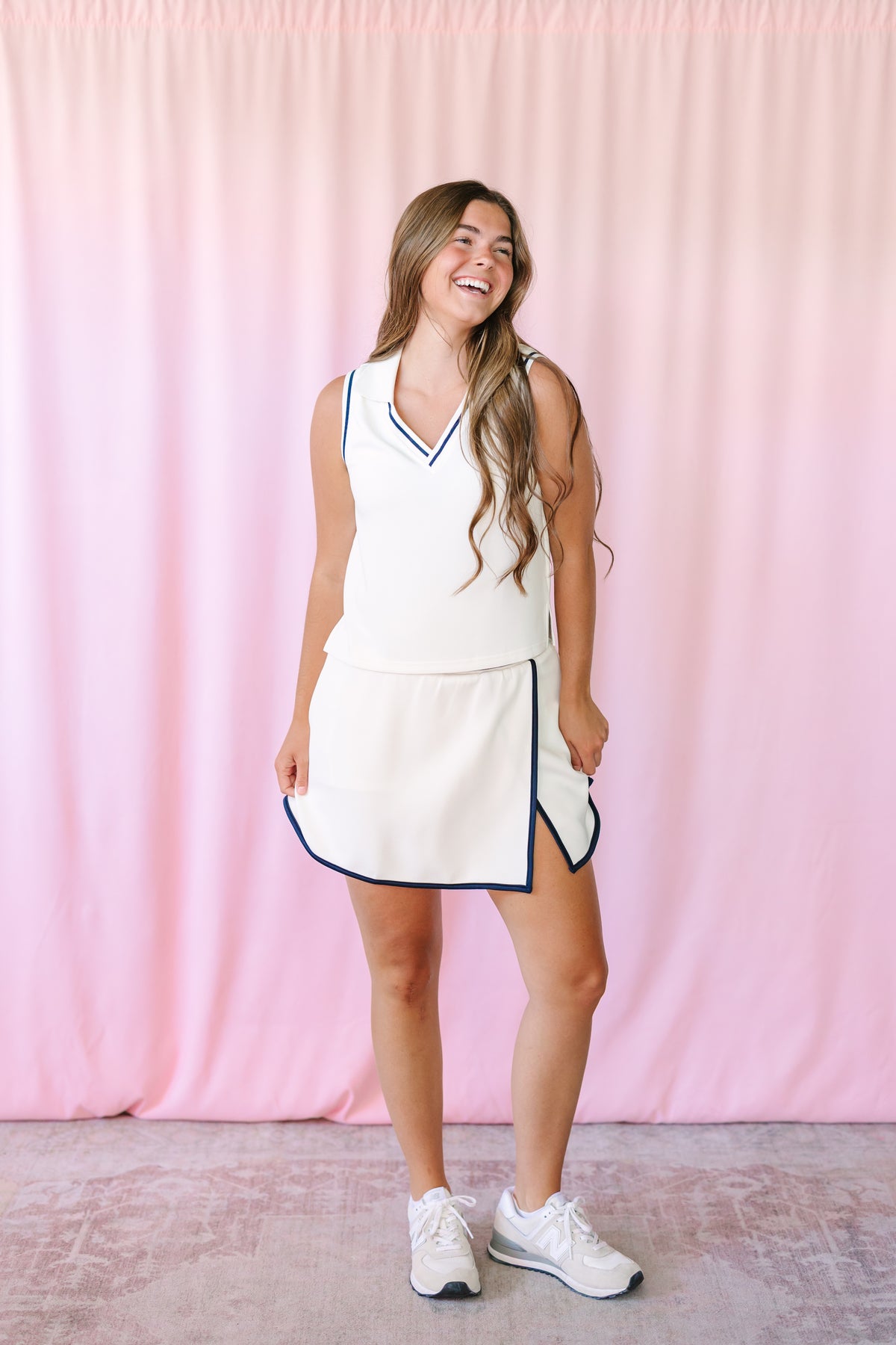 Woman laughing in a white matching set featuring a sleeveless V-neck top and mini skirt with navy tipping trim and side slit, styled with white sneakers