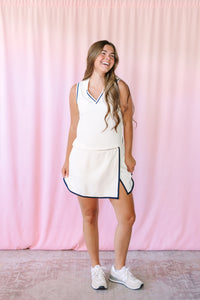 Woman laughing in a white matching set featuring a sleeveless V-neck top and mini skirt with navy tipping trim and side slit, styled with white sneakers