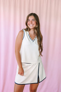 Woman smiling in a white tennis-style coordinating set with navy tipping trim — sleeveless V-neck top and mini skort with side slit, pink backdrop