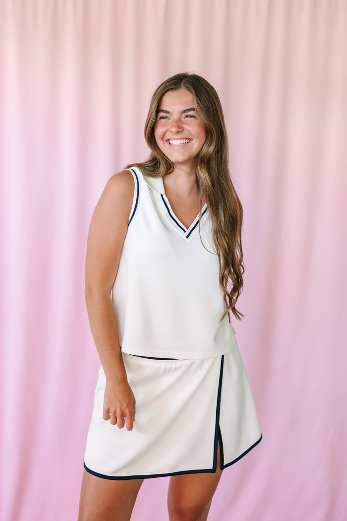 Woman smiling in a white tennis-style coordinating set with navy tipping trim — sleeveless V-neck top and mini skort with side slit, pink backdrop
