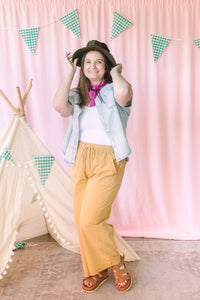 woman in golden yellow wide-leg pants styled with a white tee, denim shirt, and black hat, accessorized with a pink bandana
