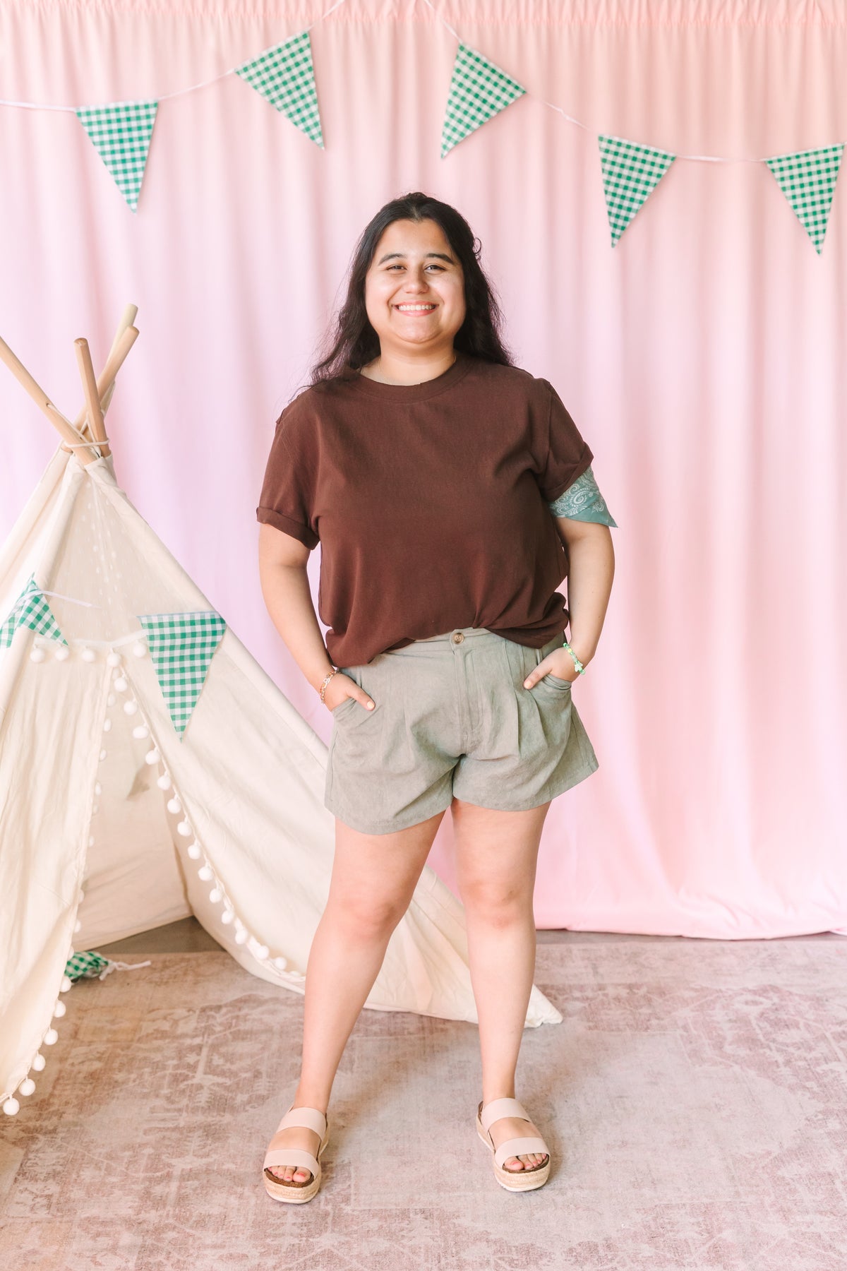 Woman smiling in a chocolate brown crew neck tee and sage green pleated high-waist shorts styled with tan platform sandals, pink backdrop with green gingham pennant banner and white teepee