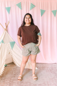 Woman smiling in a chocolate brown crew neck tee and sage green pleated high-waist shorts styled with tan platform sandals, pink backdrop with green gingham pennant banner and white teepee