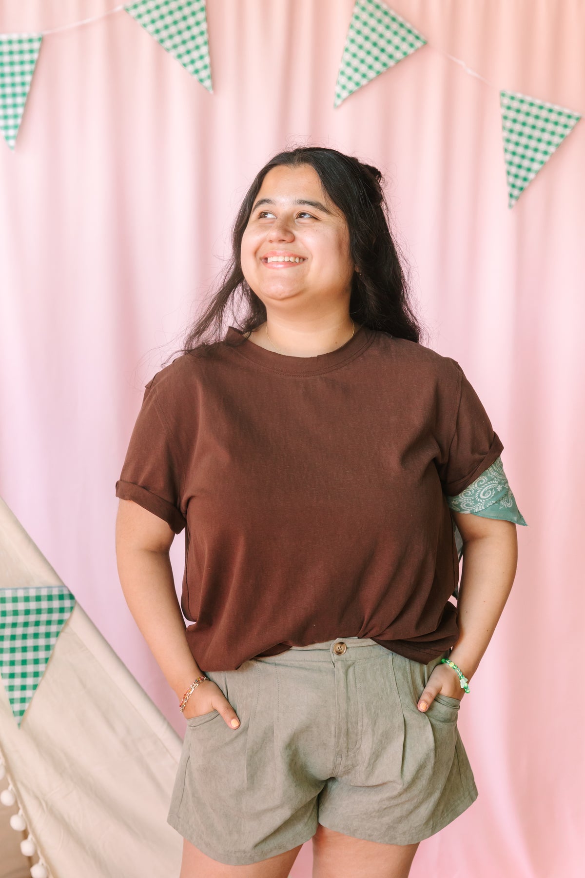 Woman smiling in a chocolate brown short-sleeve crew neck tee with rolled cuffs and sage green pleated high-waist shorts, styled with a teal paisley bandana on sleeve, pink backdrop with green gingham pennant banner