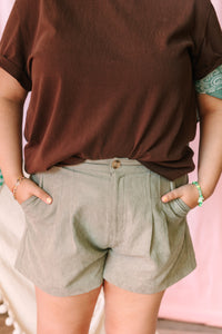 Close-up of sage green pleated high-waist shorts with single button closure and side pockets, paired with a chocolate brown tee