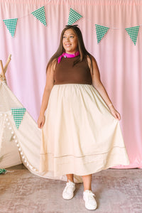 Woman in a brown sleeveless top and cream tiered midi skirt with ladder lace hem detail, styled with a pink neck scarf and white sneakers, standing in front of a pink curtain backdrop with green gingham pennant flags and a white teepee