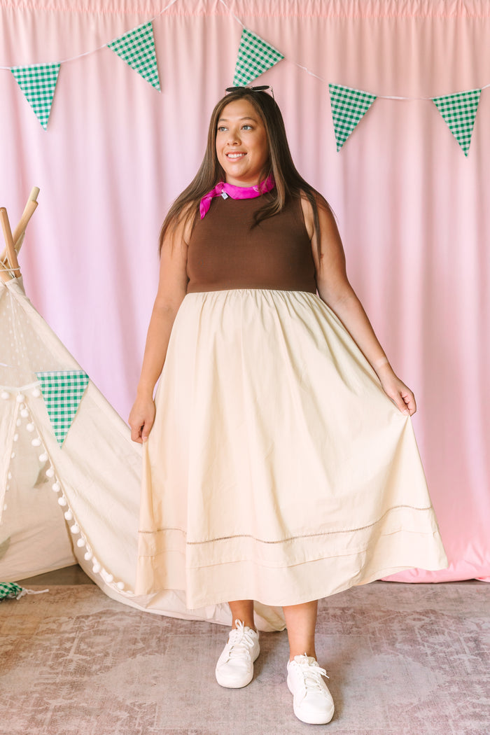 Woman in a brown sleeveless top and cream tiered midi skirt with ladder lace hem detail, styled with a pink neck scarf and white sneakers, standing in front of a pink curtain backdrop with green gingham pennant flags and a white teepee