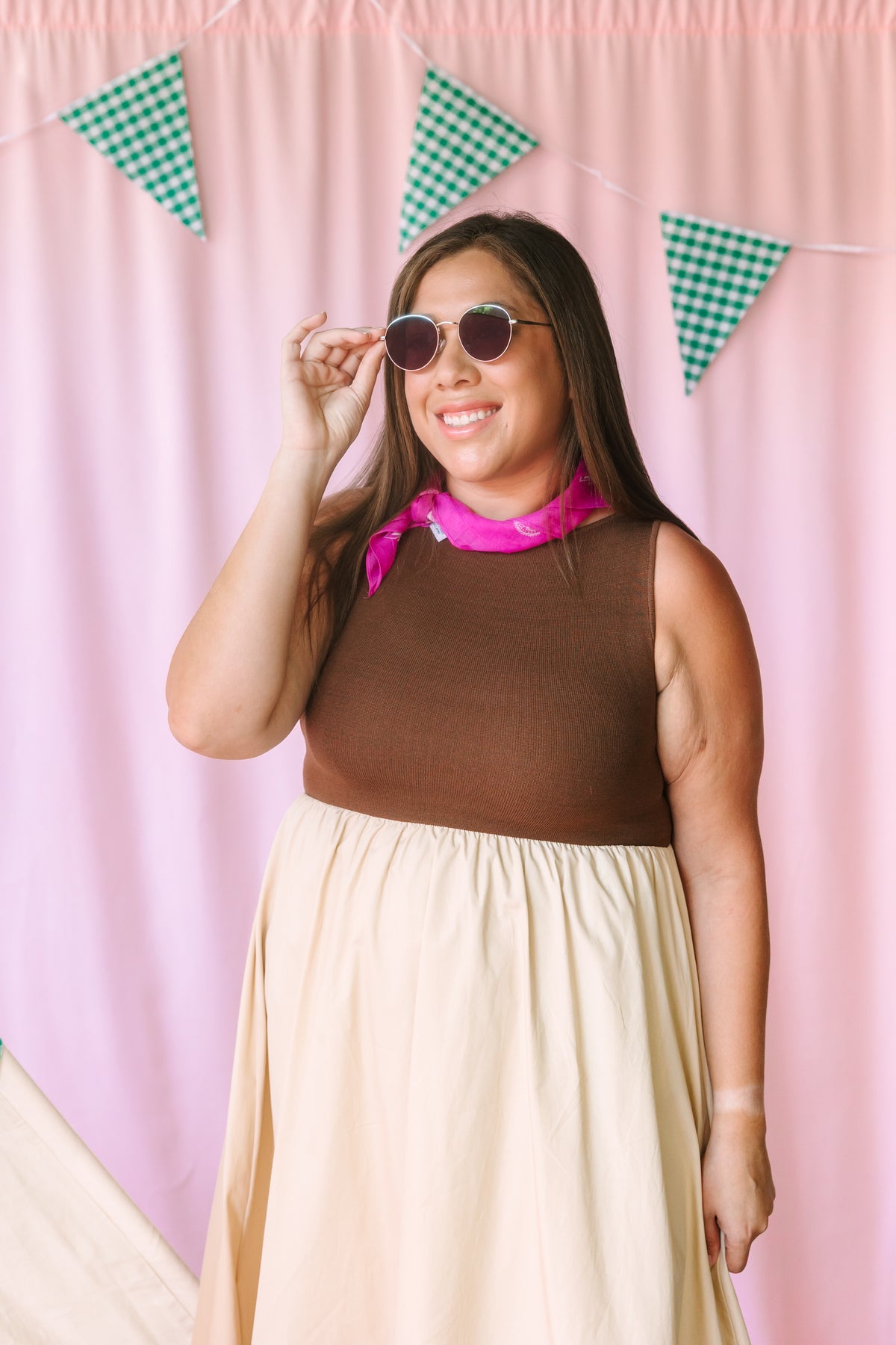 Woman smiling and holding round sunglasses, wearing a brown sleeveless ribbed top with a cream midi skirt and pink neck scarf, against a pink backdrop with green gingham pennant flags