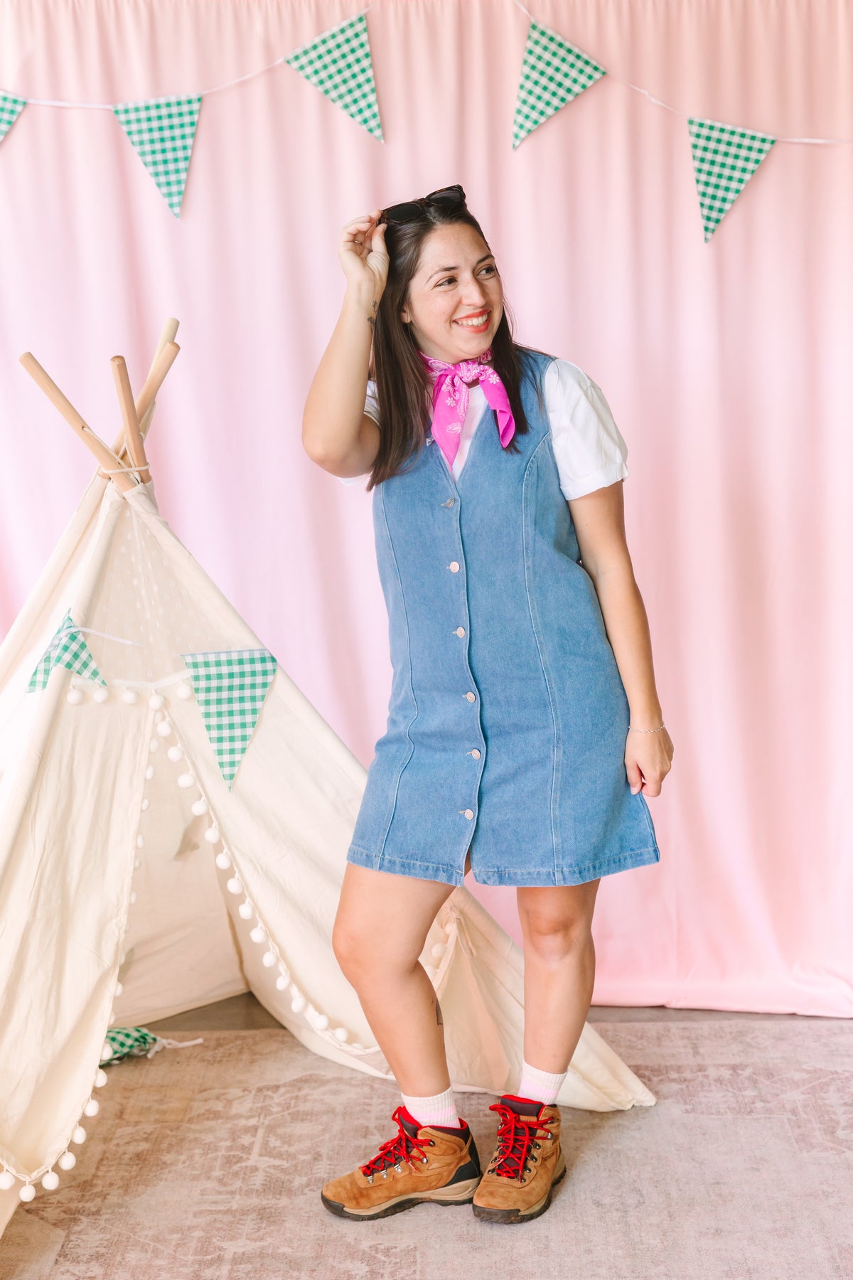 Woman wearing a light wash V-neck button-front denim dress over a white tee, styled with a pink paisley bandana, white socks, and brown lace-up hiking boots, posing in front of a pink backdrop with a white teepee